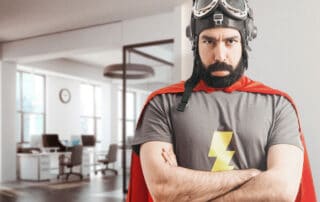 Foto de um escritório, no foco está um homem fantasiado de herói com expressão de bravo, aparentando descontentamento por trabalhar no carnaval.