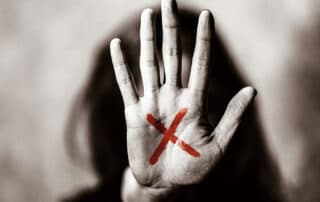 Ilustrando a capa da matéria “O que significa um X vermelho na mão?” exibimos uma fotografia monocromática, preto e branco, de uma mulher. Em primeiro plano temos a mão dela aberta em direção a camêra, em sua palma vemos um X desenhado na cor vermelho, no plano de fundo, desfocado, é possível ver seus longos cabelos escuros, o rosto está encoberto pela palma do primeiro plano.