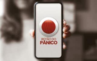 Ilustrando a capa da matéria “Botão do pânico, tecnologia aliada de mulheres vítimas de violência” exibimos uma fotografia. No primeiro plano temos a mão de uma mulher exibindo a tela do seu smartphone, em tal tela vemos uma ilustração de um grande botão vermelho. No plano de fundo, bastante desfocado e em preto e branco, vemos sem muitos detalhes, a mulher que exibe seu smartphone.