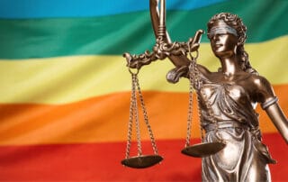 Fotografia colorida ilustra a matéria "STJ: Lei Maria da Penha pode ser aplicada para mulheres transexuais". Na imagem vemos uma estátua de bronze da Thêmis, a deusa da justiça, em primeiro plano e no plano de fundo, a bandeira LGBTQ+.