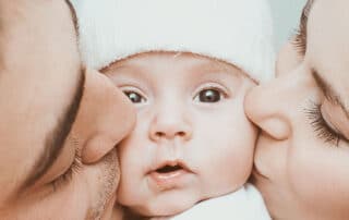 Ilustrando a matéria "Licença-maternidade x licença parental", temos uma fotografia colorida onde é possível ver, do lado esquerdo da tela, o rosto de um homem que está beijando a bochecha de um bebê localizado no meio da tela, já do lado direito, temos uma mulher, ela também está beijando a bochecha do bebê