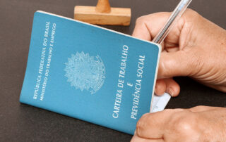 Fotografia colorida ilustra a capa da matéria "Saque do FGTS 2022 começa nesta quarta 20/04/22, veja calendário". Vemos a mão de alguém preenchendo uma Carteira de Trabalho e Previdência Social sobre uma superfície preta. Ao fundo da fotografia temos um carimbo de madeira.