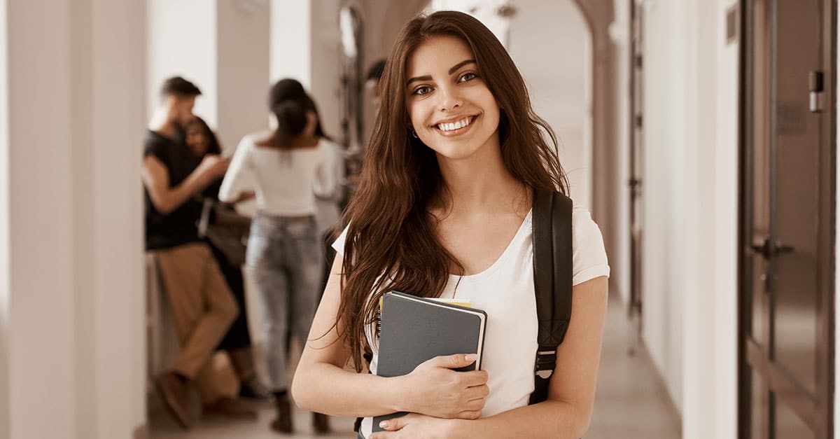 Ilustrando a capa da matéria "Faculdade de Direito: 7 coisas que a faculdade não te ensina", temos uma jovem mulher, ela está sorrindo, segura dois cadernos e apoiado ao seu ombro vemos uma mochila.