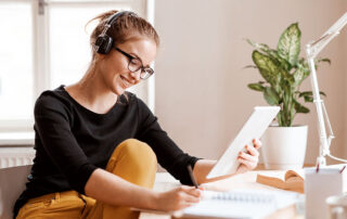 Ilustrando a capa da matéria autoral "Como se preparar para a OAB: 8 canais gratuitos", temos uma jovem mulher sentada casualmente. Ela está de fone de ouvidos sem fio, segura um tablet e acima de sua mesa de estudo, conferimos cadernos e folhas.
