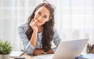 Como imagem de capa da matéria 9 dicas para advogados iniciantes, temos uma jovem mulher sentada olhando para a direção da câmera e sorrindo. A sua frente vemos um laptop e alguns papéis.