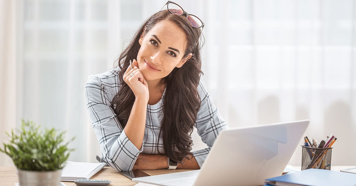 Como imagem de capa da matéria 9 dicas para advogados iniciantes, temos uma jovem mulher sentada olhando para a direção da câmera e sorrindo. A sua frente vemos um laptop e alguns papéis.