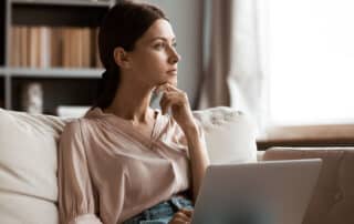 Ilustrando a capa da matéria - Como escolher uma pós-graduação em Direito – conferimos a fotografia de uma jovem mulher sentada em um sofá com seu laptop apoiado no colo. A mulher está olhando para o lado, com uma das mãos no seu queixo, seu semblante é de uma pessoa que está pensando.