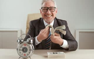 Como capa da matéria "Revisão de aposentadoria: Aposentado passou a receber mais que o dobro", vemos um homem mais velho, de óculos, sentado em uma mesa e segurando notas de dinheiro na mão. Também vemos um relógio sobre a mesa.