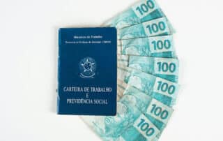 Como capa da matéria “Correção do saldo do PIS/PASEP. Como funciona?”, vemos uma carteira de trabalho brasileira com notas de 100 reais ao fundo.