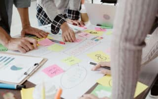 Como capa da matéria “O que é e como funciona o Legal Design”, vemos um grupo de pessoas discutindo ideias ao redor de uma mesa cheia de papéis e anotações em um escritório.