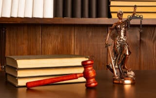 Como capa da matéria “Livros para advogados – Parte 2”, temos livros sobre uma mesa com um martelo de juiz e uma estátua da Justiça.