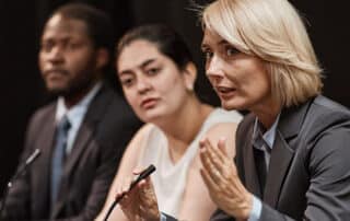 Como capa da matéria “Dicas de oratória para advogados iniciantes”, vemos a imagem de uma advogada fazendo uma apresentação diante de outras pessoas.